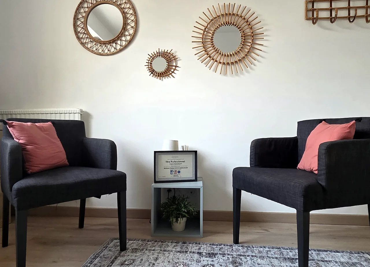 Deux fauteuils gris face à face dans le bureau de Christel Louvet, trois miroirs ronds en rotin au mur et un diplôme encadré posé sur une sellette